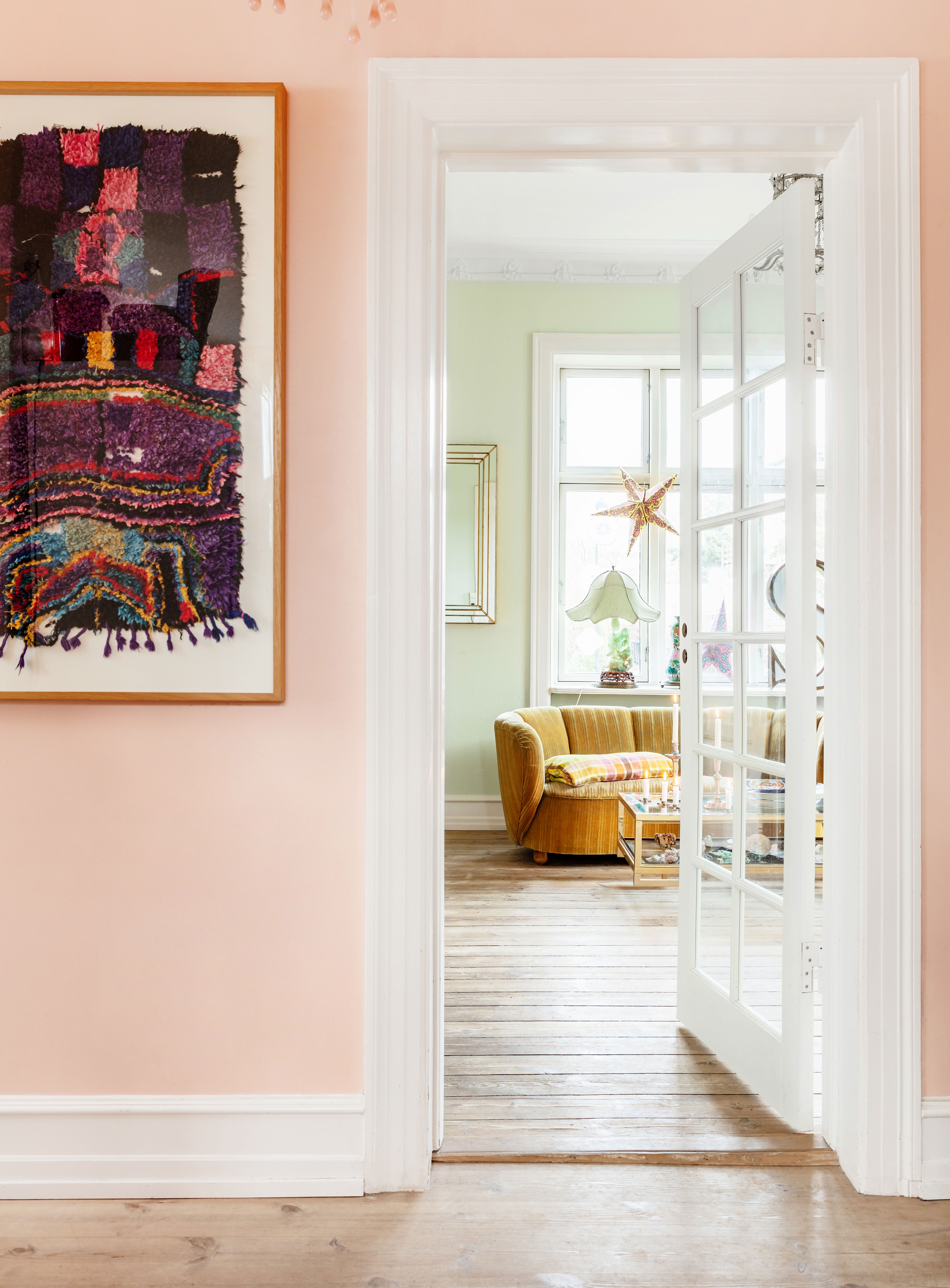 Et fint kig fra den rosa spisestue ind til stuen, der er malet i en sart æblegrøn farve. Den gule banansofa har Shiri købt på auktion, ligesom den kinesiske antikke Guanyin-lampe. Spejlet er et antikt spejl fra Belgien, som minder Shiri om hendes logo fra Carré. Det indrammede marokkanske tæppe, har Shiri fået af sin bror, kunstneren Tal R.